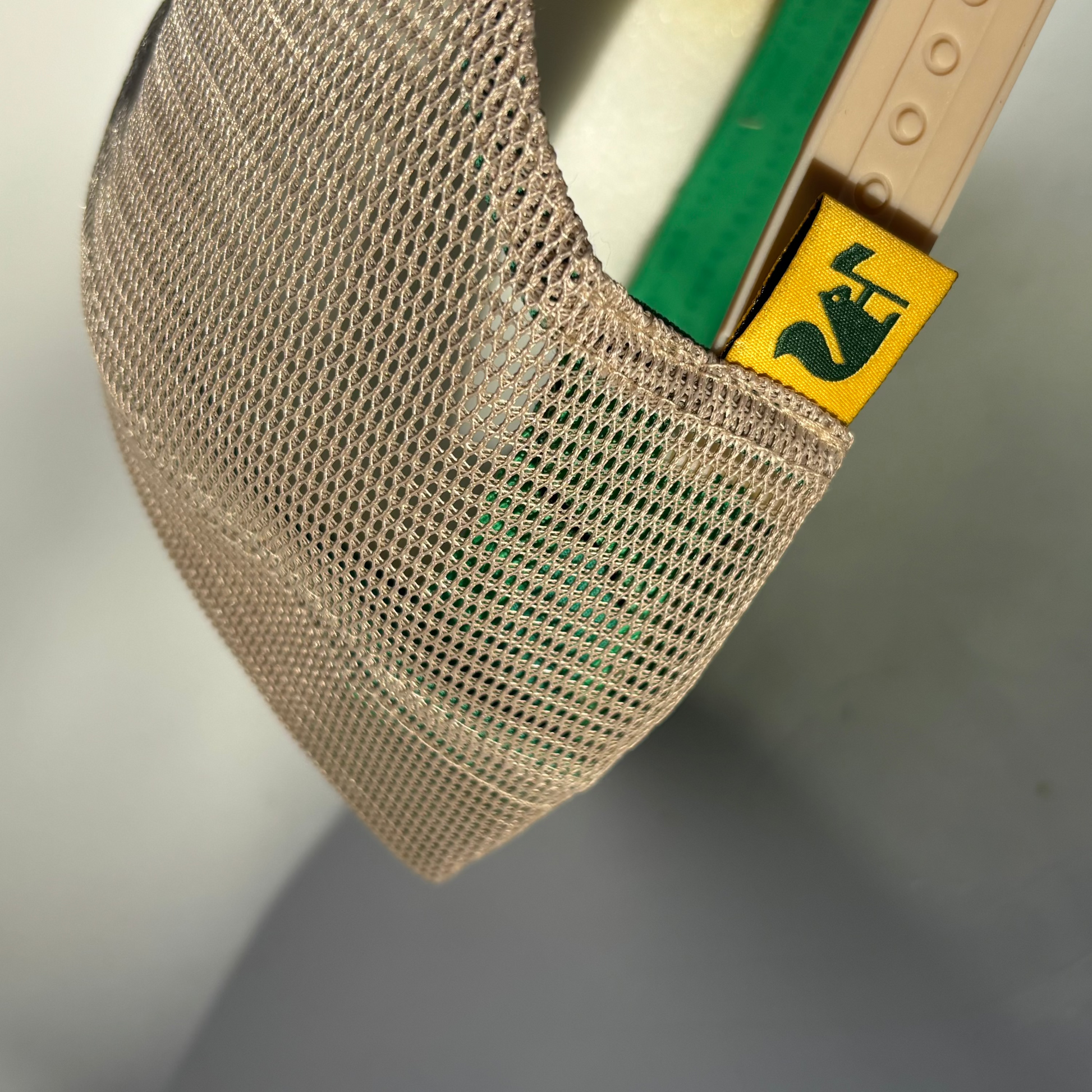 Close-up of a beige mesh cap with a green strap and yellow label on a neutral background